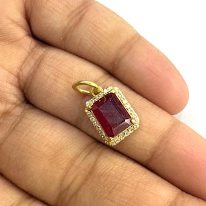 Puede incluir: Colgante de oro con una piedra preciosa rectangular de color rojo intenso en el centro. La piedra está rodeada por un borde de pequeñas piedras claras. El colgante tiene un pequeño lazo en la parte superior para una cadena.