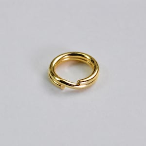 May include: A gold-colored metal jump ring featuring a double-loop design. The circular component has a smooth, reflective surface and a split opening, constructed from a single piece of wire coiled to create two overlapping circular layers.