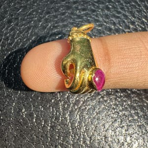 Pode incluir: Um pingente dourado em forma de mão, com uma pedra preciosa vermelha. O pingente tem uma argola para uma corrente. O fundo é uma superfície escura e texturizada. O pingente é provavelmente de ouro.