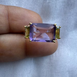 以下が含まれることがあります： 中央に大きな長方形のファセットカットが施された紫色の宝石を配し、その両側に小さな長方形のファセットカットが施された黄緑色の宝石を添えたゴールドリング。すべて細いゴールドバンドの爪留めセッティング。