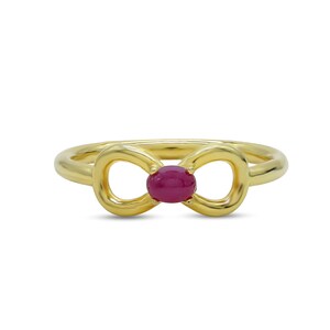 Könnte beinhalten: Ein goldfarbener Ring mit einem bogenförmigen Design. Der Ring hat einen zentralen, oval geformten, tiefroten Edelstein. Das Band des Rings ist glatt und poliert und schafft eine schlichte, aber elegante Ästhetik. Der Ring ist vor einem weißen Hintergrund platziert.