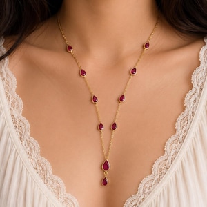 Puede incluir: Un collar de cadena dorada con múltiples piedras preciosas de color rojo intenso en forma de lágrima, espaciadas uniformemente. El collar termina en un diseño de caída vertical con dos piedras rojas en forma de lágrima apiladas en el centro.