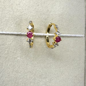 May include: A pair of gold hoop earrings, each with a central red gemstone and smaller clear stones. The earrings are presented on a white string against a neutral backdrop. The earrings are a classic jewellery item.
