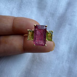 Peut inclure: Une bague en or ornée d'une grande pierre précieuse rectangulaire rose foncé avec des inclusions internes visibles, flanquée de deux pierres rectangulaires vert clair de chaque côté, le tout monté sur un anneau en or avec des griffes.