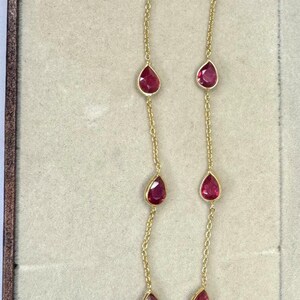 Pode incluir: Um delicado colar de ouro com pedras preciosas vermelhas em forma de gota. As pedras preciosas são espaçadas ao longo de uma fina corrente de ouro, criando um design sofisticado e elegante. O colar é exibido em um fundo neutro.