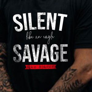 Può includere: Una t-shirt nera con testo bianco che recita SILENT, seguito dal testo in corsivo like an eagle, poi la parola SAVAGE in un carattere bianco invecchiato e infine il testo like a wolf all'interno di un banner rosso in fondo alla grafica.
