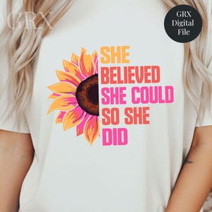 Può includere: T-shirt bianca con una grafica di girasole colorato sul lato sinistro. Il testo "SHE BELIEVED SHE COULD SO SHE DID" è stampato in arancione, rosso e rosa. Un logo di file digitale è nell'angolo in alto a destra.