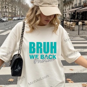 Puede incluir: Camiseta color crema con el texto "BRUH WE BACK Protos" en turquesa y negro. La persona lleva una gorra de béisbol beige y un bolso de hombro negro acolchado. El fondo muestra una calle de la ciudad.