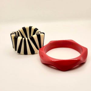 May include: A black and white striped stretch bracelet composed of rectangular segments alongside a solid red bangle with a faceted, hexagonal outer edge. Both bracelets feature a smooth, glossy finish and are presented together as a set.
