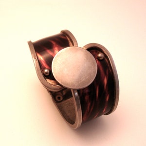 May include: A silver and brown metal bracelet with a round, silver, decorative element. The bracelet has a textured, brown surface.