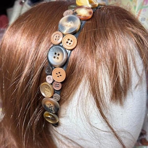 May include: A black headband decorated with a variety of vintage buttons in shades of brown, tan, and cream. The buttons vary in size and style, creating a unique and textured accessory. The headband is a stylish hair accessory.