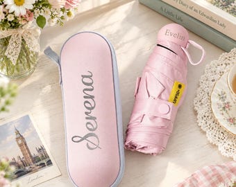 May include: A light pink collapsible umbrella and a matching hard-shell carrying case. The case features the name Serena in cursive script. The umbrella handle is personalised with the name Evelin and includes a yellow UPF50+ tag.