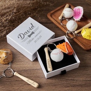 May include: A white gift box with the text David, Make contact!, and 26.09.28 printed on the lid. Inside the box is a keychain with a miniature baseball, a baseball bat engraved with Chantal, and an orange glove. Other keychains are nearby.