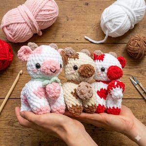 May include: Three handmade crochet cow plushies in white, pink, and brown. The plushies have small horns and smiling faces. The background includes yarn balls in pink, white, and red, along with a crochet hook and scissors.