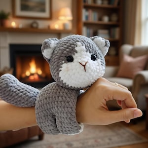 May include: A gray and white crocheted cat wrist cuff. The cat has black eyes and a brown nose. The tail wraps around the wrist. The background includes a fireplace and a cozy living room setting.