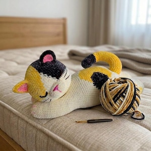 May include: A handmade crochet cat in cream, yellow, black, and pink, lying on a bed. A ball of yarn and a crochet hook are next to the cat. The cat has a yellow and black head, pink ears, and a black tail.