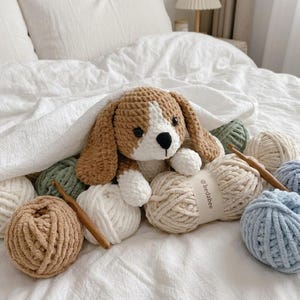 May include: A handmade crochet dog toy in brown, white, and black, nestled among various colored yarn balls and crochet hooks. The dog is under a white blanket on a bed. The yarn colors include beige, green, and blue.