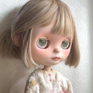 May include: A doll with a large head, green eyes, and light brown hair. The doll has a pale complexion with rosy cheeks and a small mouth. It wears a white and pink patterned top with small pink buttons.