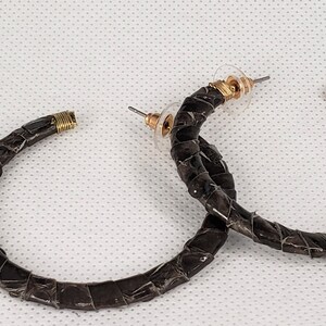 以下が含まれることがあります： 編み込みのデザインが施された、黒いフープイヤリングのペア。イヤリングは革製で、金色の金具が付いています。