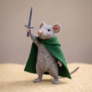 Könnte beinhalten: Handgefertigte Filzmaus-Figur, gekleidet als Krieger. Die graue Maus trägt einen grünen Umhang und hält ein kleines graues Schwert hoch. Die Maus hat eine rosa Nase und detaillierte Merkmale, die auf einer hellbraunen Oberfläche steht.