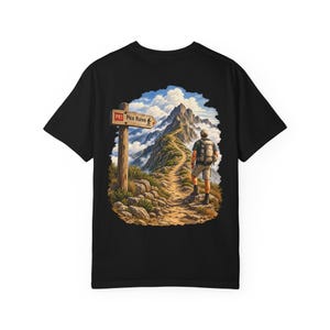 May include: A black t-shirt featuring a graphic print on the back. The illustration depicts a hiker with a backpack standing on a rocky mountain trail. A wooden signpost points toward Pico Ruivo, set against a backdrop of jagged peaks and clouds.