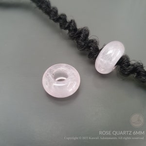 May include: Two pink rose quartz donut beads, each with a hole in the center. The beads are approximately 6mm in diameter. The beads are strung on a black cord.
