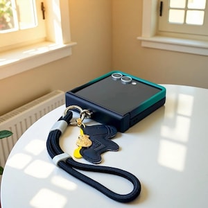 May include: A navy blue and teal smartphone case with a matching wrist strap and charm. The case has a teal border and two camera lenses. The wrist strap is black with gray accents, and the charm is a navy blue boot with yellow and gold details.