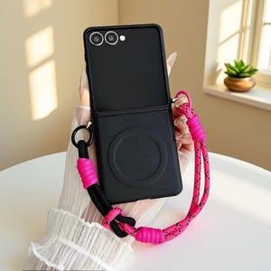 Puede incluir: Funda de teléfono negra con un cordón rosa y negro. La funda del teléfono está abierta, revelando las lentes de la cámara. El cordón está hecho de un material similar a una cuerda rosa y negra y está sujeto a la funda con un anillo negro.