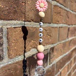 Könnte beinhalten: Ein Perlenornament mit dem Namen "LINA" in weißen Buchstabenperlen. Es zeigt eine rosa Gänseblümchen, Holzperlen, rosa Perlen und einen durchsichtigen Kristallkugel. Hängt vor einer Backsteinmauer.