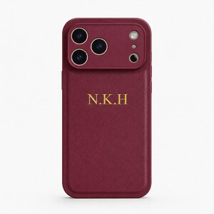 Peut inclure: Une coque de smartphone bordeaux profond à la finition texturée, dotée d'un module photo surélevé et des initiales N.K.H dorées en relief au centre. Le design comprend trois ouvertures circulaires pour les objectifs et une ouverture flash.