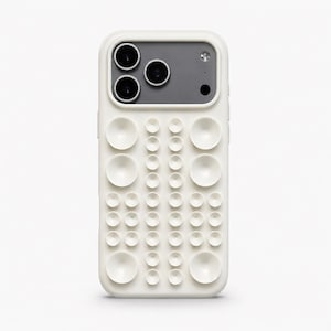 Peut inclure: Coque de téléphone blanc cassé avec un design texturé unique. La coque présente une série d'éléments circulaires en relief de différentes tailles, créant une surface tactile. La zone de la caméra est grise avec trois objectifs noirs et un flash.