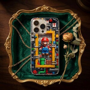 May include: Clear phone case featuring a colorful Mario figure and a yellow and red brick design. The case has a transparent back, showcasing the phone's camera and design. The case is displayed on a green velvet surface with a gold frame and a dried flower.