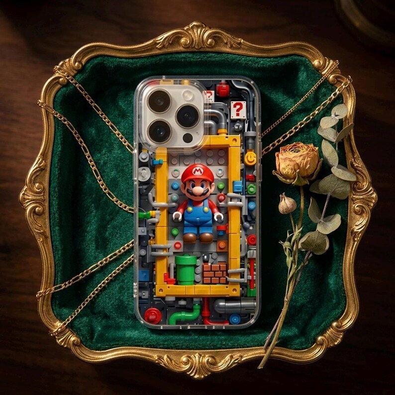 May include: Clear phone case featuring a colorful Mario figure and a yellow and red brick design. The case has a transparent back, showcasing the phone's camera and design. The case is displayed on a green velvet surface with a gold frame and a dried flower.