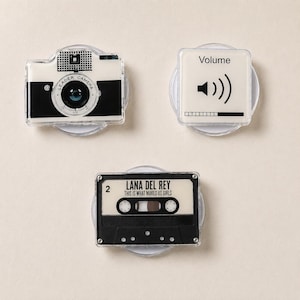 May include: Three circular phone grips featuring distinct designs: a black and white vintage-style camera labeled Leader Camera, a white square with the text Volume and a speaker icon, and a black cassette tape labeled Lana Del Rey This Is What Makes Us Girls.