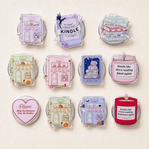 May include: A collection of eleven acrylic pins featuring book-themed illustrations, including storefronts, stacks of books, candles with text, and heart-shaped designs. The pins display various colours like pastel purple, pink, green, and red with printed text.