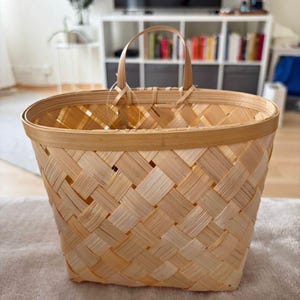 May include: A woven basket with a handle, crafted from light-colored wood strips. The basket has an oval shape and a textured, crisscross pattern. The handle is attached with small wooden pieces. The basket is empty and sits on a neutral-colored surface.