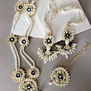 May include: A full jewellery set comprising a necklace, earrings, bracelet, and headpiece. The set features white pearl strands and gold-toned floral accents with black and green centres. The jewellery is displayed on a neutral surface.