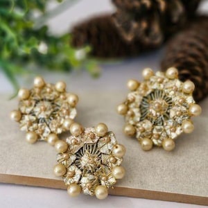 May include: Three gold-toned, floral-shaped earrings with pearl accents. The earrings have a detailed, textured design with small, round pearls around the edges. The earrings are on a light-colored surface.