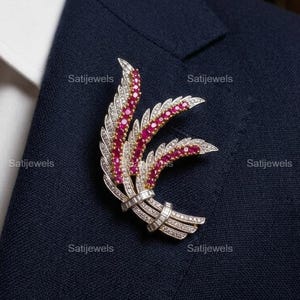 May include: A silver and ruby brooch in a leaf design, set against a navy blue suit. The brooch features a central red section with white accents and is secured with a pin. The suit has a white collared shirt underneath.