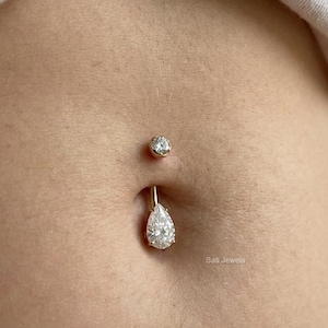 May include: A belly button piercing set featuring a small round stud and a pear-shaped dangle. Both pieces are gold-toned and adorned with clear, sparkling gemstones. The jewelry is displayed against a skin tone background.