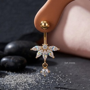 May include: A gold-colored belly button ring featuring a floral design with clear, sparkling gemstones. The ring has a dangling charm with a single gemstone. The background is dark with black stones and sand. The text "Sati Jewels" is visible.