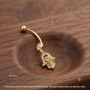 May include: A gold-toned curved barbell belly button ring featuring a small clear round gemstone and a dangling Hamsa hand charm encrusted with tiny clear stones. The text SATI JEWELS is visible in the lower left corner of the image.