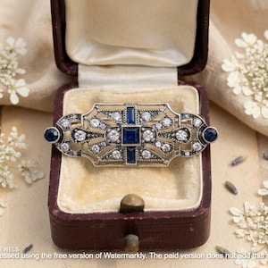 Könnte beinhalten: Eine rechteckige silberfarbene Brosche mit einem zentralen quadratischen blauen Stein, vier umgebenden rechteckigen blauen Steinen und zwei runden blauen Steinen an den Enden. Der Metallrahmen ist mit zahlreichen kleinen klaren Steinen verziert.