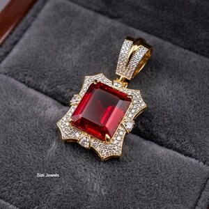 May include: A gold pendant featuring a large, rectangular, deep red gemstone at the center, surrounded by a border of small, clear, pavé-set stones. The bail is also encrusted with clear stones. The text Sati Jewels is visible on the left.