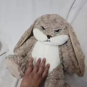 May include: A plush bunny with a stern expression. The stuffed animal has a light brown, textured body, long ears, and a white belly. It features red eyes and a black frown. The bunny is lying on a white surface.
