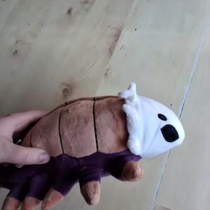 May include: A plush toy resembling a woodlouse. It features a brown, segmented back, a white head with black eyes, and purple legs with orange tips. The toy is made of soft fabric.
