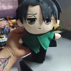 Peut inclure: Poupée en peluche avec des cheveux noirs et une cape verte. La poupée a une expression neutre et porte une tenue noire. La poupée est tenue dans une main.
