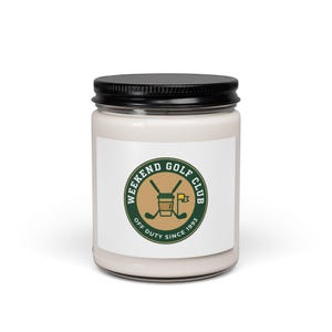 May include: A white candle in a glass jar with a black lid. The label features a green and brown circular logo with the text "WEEKEND GOLF CLUB OFF DUTY SINCE 1993". The logo includes crossed golf clubs, a golf flag, and a golf cart.