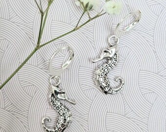 May include: A pair of silver-toned seahorse earrings featuring a detailed, textured scale pattern on the body and a curved tail. Each seahorse charm is attached to a circular hoop finding, creating a dangling design with a metallic finish.