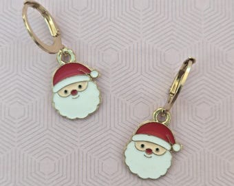 May include: A pair of gold-toned hoop earrings featuring dangling Santa Claus charms. Each charm is crafted with red and white enamel, depicting a smiling Santa face with a red hat, a white beard, and a small red nose, suspended from a simple gold hoop.